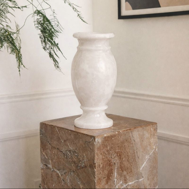 White marble vase on a marble pedestal against a wall with a framed abstract painting.